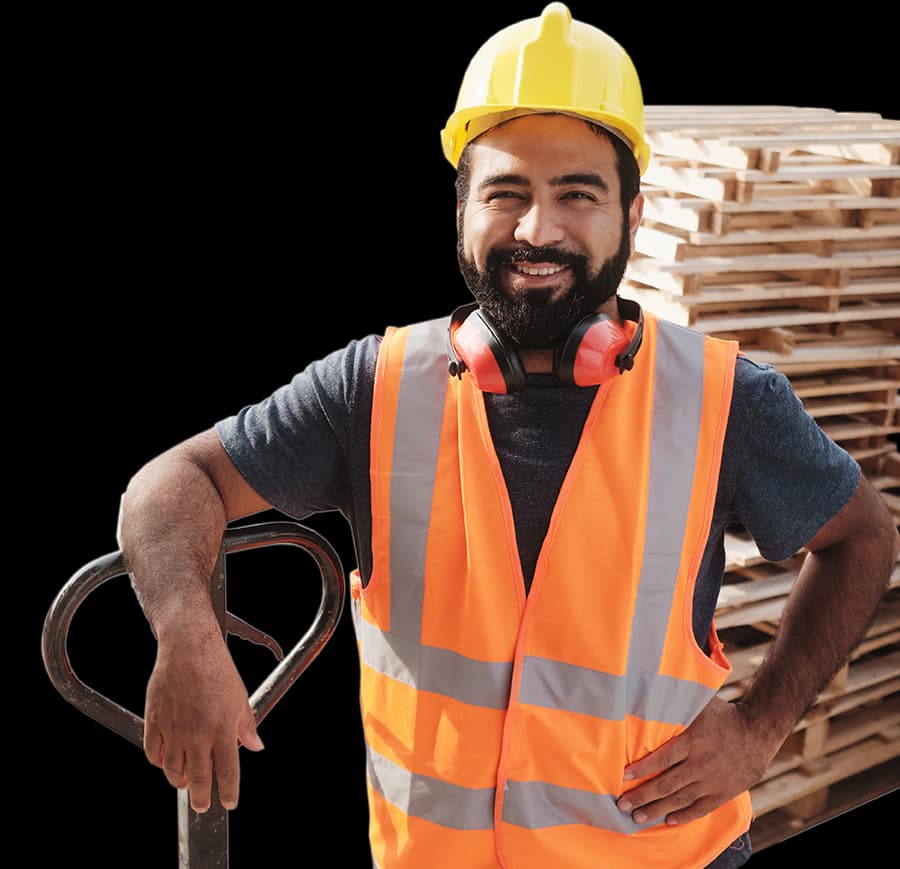 Male pallet worker wearing a safety helmet holding a pallet jack standing in front of a stack of wooden pallets in background