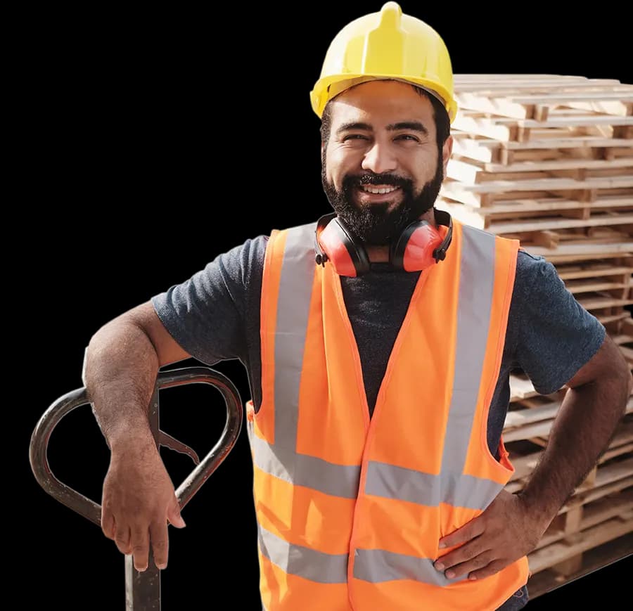Male pallet worker wearing a safety helmet holding a pallet jack standing in front of a stack of wooden pallets in background