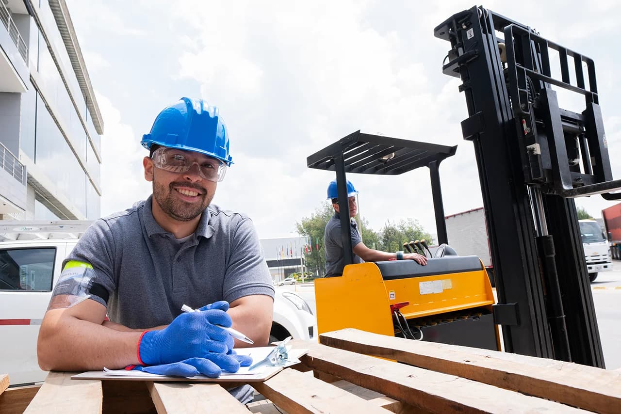 Man holding clipboard with arms resting on wood pallet and warehouse working driving forklift in background