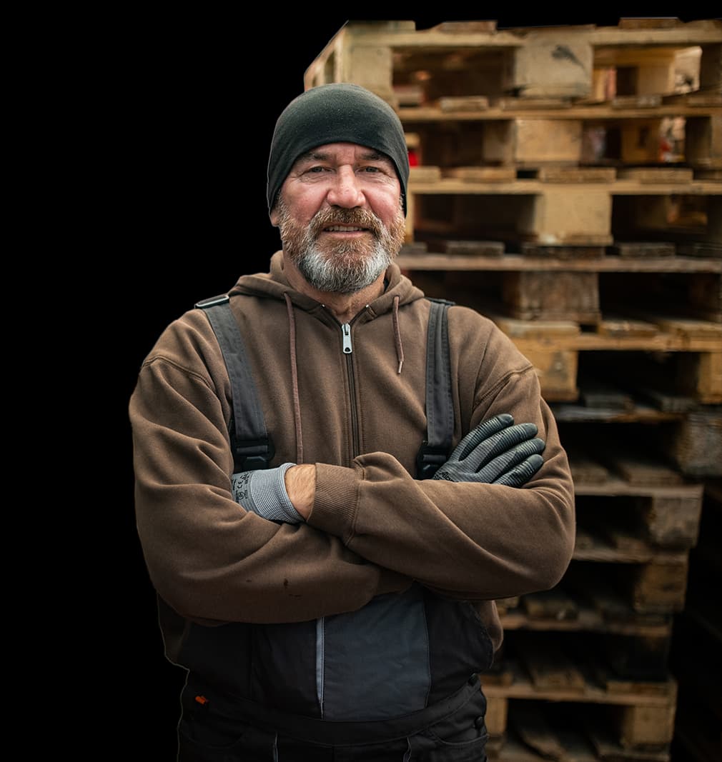 Man wearing sweater and beanie smiling and standing in front of a stack of pallets to haul-off